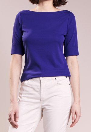 Kvinde iført en tætsiddende, kongeblå top med albuelange ærmer, kombineret med højttaljede, offwhite bukser mod en ensfarvet baggrund.