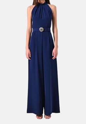 Vrouw draagt een mouwloze marineblauwe jumpsuit met halternek, een decoratieve riem en broek met wijde pijpen, staand tegen een effen achtergrond.