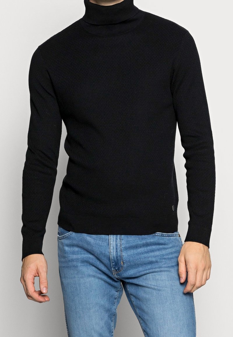 Svart polotröja gjord av texturerad stickad tyg, med långa ärmar och en åtsittande design, kombinerad med ljusblå denimjeans.