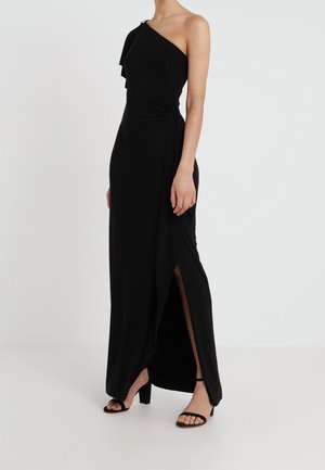 Femme portant une longue robe noire asymétrique avec une fente latérale et des sandales à talons ouvertes noires, debout devant un fond blanc uni.