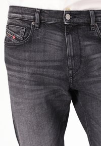 Jean en denim gris foncé avec une légère texture usée, comprenant une poche avant et un système de fermeture à bouton argenté. Accentué d'une étiquette de marque.