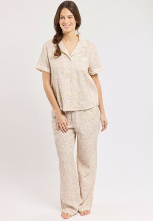 Femme aux longs cheveux bruns en tresse lâche porte un haut de pyjama à manches courtes à motifs floraux pâles et un pantalon assorti, debout pieds nus sur un fond uni.