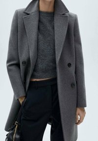 Gray wool coat with a structured fit, notched lapels, double-breasted closure, and side pocket. Worn over a dark sweater and striped pants.