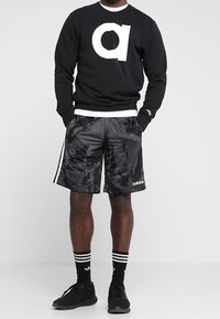 Maglione nero con un grande logo bianco "a", abbinato a pantaloni corti neri in camouflage con strisce bianche. Scarpe da ginnastica nere e calzini a righe.