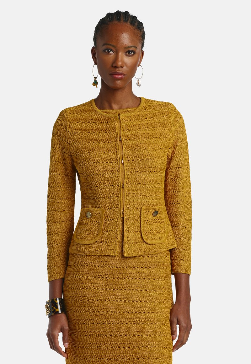 Frau mit geflochtenem Haar, die eine senfgelbe, strukturierte Strickjacke mit Knöpfen und passenden Rock trägt, dazu goldene Creolen-Ohrringe und ein Armreif.