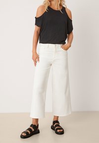 Femme portant un haut noir à épaules dénudées, un pantalon blanc large et court, et des sandales noires à lanières, se tenant devant un fond uni.