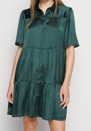 Kvinde iført en kortærmet, mørkegrøn, satin kjole med knapper og lagdelte, rynkede nederdel.