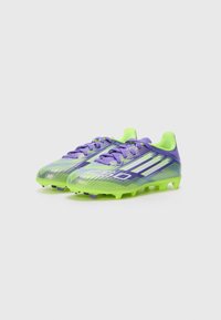Des chaussures de football violettes et vert néon avec une tige synthétique texturée, un système de laçage et des rayures blanches proéminentes sur les côtés.