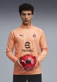 Športový dres svetloružovej farby s žltým geometrickým vzorom, zipsovým golierom a logom na hrudi. Model drží červenú futbalovú loptu s čiernymi akcentmi.