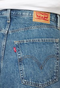 Poche arrière d'un jean Levi's bleu avec une étiquette Levi's rouge et un patch en cuir de marque sur la ceinture, porté avec une chemise blanche.