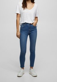 Jeans ajustados de mezclilla azul con una cintura alta, combinados con una camiseta blanca de manga corta y zapatillas blancas. Diseño simple y casual.