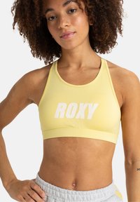 Žltá športová podprsenka s bielym logom "ROXY". Vyrobená z hladkej látky, s pretekárskym strihom a podporným pásom pod prsiami.