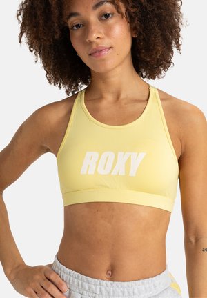 Žltá športová podprsenka s bielym logom "ROXY". Vyrobená z hladkej látky, s pretekárskym strihom a podporným pásom pod prsiami.