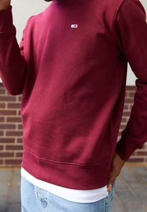 Personne portant un sweat-shirt grenat sur une chemise blanche et un jean bleu clair, debout près d'un mur en briques.