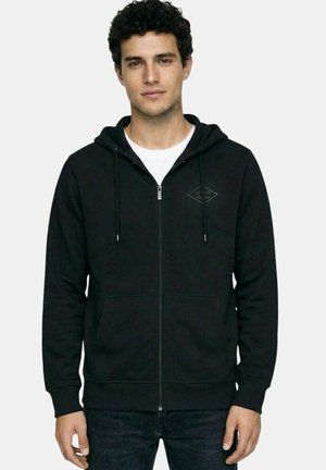 Jeune homme portant un sweat à capuche noir zippé avec des poches frontales et un t-shirt blanc en dessous, se tenant devant un fond uni.