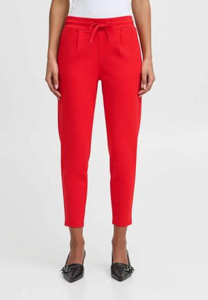 Pantalons rouges fuselés avec taille à cordon, comportant des plis avant et deux poches latérales. Tissu lisse associé à des chaussures noires pointues.