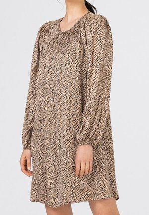 Kobieta w sukience z długim rękawem sięgającej kolan z brązowo-beżowym abstrakcyjnym wzorem w kropki, stojąca na jednolitym jasnoszarym tle.