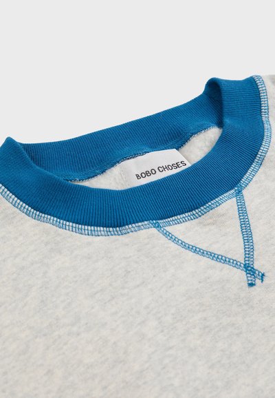 Svijetlosiva majica s plavim rebrastim izrezom, s kontrastnim plavim šavovima i etiketom s natpisom "BOBO CHOSES" na unutarnjoj strani.
