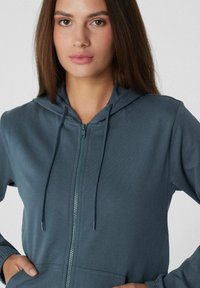 Hérmine zippée de couleur bleu foncé, avec une capuche, des cordons de serrage, une fermeture éclair en métal et des poches latérales. Fabriquée en tissu doux et texturé.