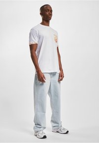 White cotton t-shirt featuring a multicolored geometric design on the sleeves, paired with light blue baggy jeans and gray sneakers.