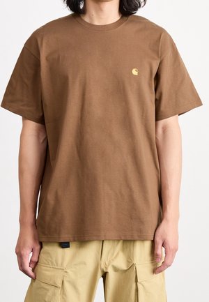 Persona che indossa una t-shirt Carhartt marrone a maniche corte con un piccolo logo giallo sul petto e pantaloni cargo beige con tasche.