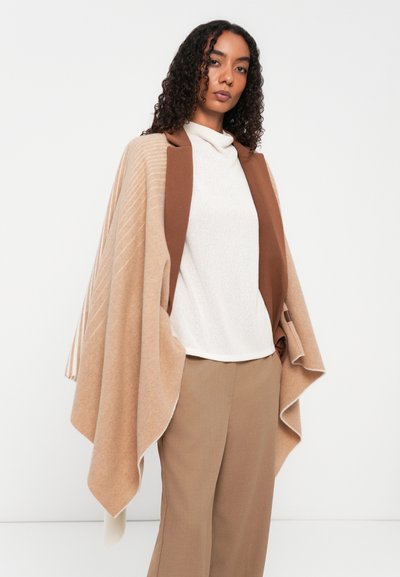 Beige strikket poncho med brun krage, med diagonale striper. Kombinert med en hvit høyhalset genser og beige bukser for et lagdelt utseende.