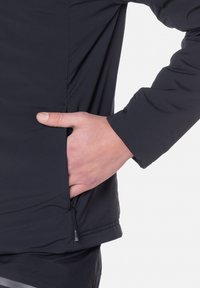 Zwarte, lichte jas gemaakt van zacht, waterafstotend materiaal. Voorzien van een zijzak met een ritssluiting en elastische manchetten.