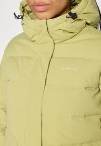 Pufota parkas gaiši zaļā krāsā ar augstu apkakli, ar izšūtām detaļām un izšūtu "sniega virsotnes" logo uz krūškurvja.