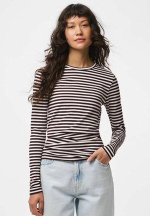 Femme aux longs cheveux foncés portant un haut à manches longues rayé noir et blanc et un jean bleu clair, une main dans la poche.