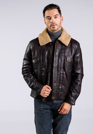Braune Lederjacke mit einem beigen Schafslederkragen; verfügt über zwei Fronttaschen, eine Knopfleiste und eine strukturierte Oberfläche. Getragen über einem grauen Rollkragenpullover.