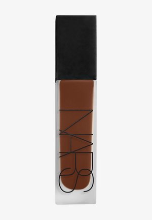 Flacon en verre rectangulaire avec un fond de teint liquide brun foncé et un bouchon carré noir, étiqueté avec un texte noir vertical.