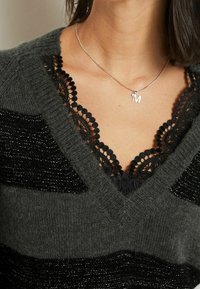 Pull en tricot gris avec des accents en dentelle noire, doté d'un décolleté en V et de rayures texturées. Collier en argent avec un pendentif en forme de lettre "M".