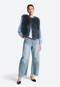 Grijze faux bontvest, gedragen over een lichtblauw lange-mouwen blouse, gecombineerd met wijd uitlopende lichtblauwe jeans met slijtage en puntige zwarte hakken.