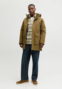 Brun vandtæt jakke med hætte, to for-lygter, lagd ovenpå en ternet skjorte. Båret med løse blå jeans og brune sko.