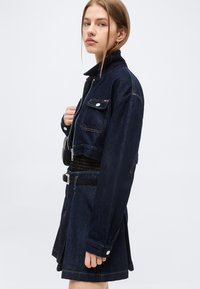 Νέα γυναίκα με ανοιχτό καστανά μαλλιά, φοράει σκούρο μπλε κοντό τζιν μπουφάν και ταιριαστή φούστα, μπροστά σε απλό φόντο.