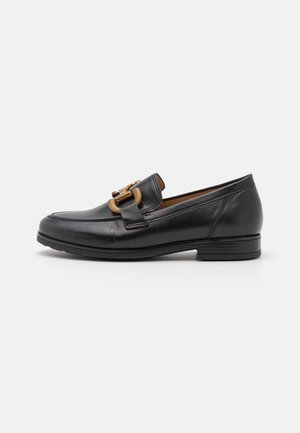 Mocasín de cuero negro con tacón bajo y hebilla decorativa de tono dorado en la parte superior, mostrado de perfil sobre fondo blanco.