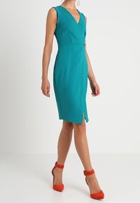 Tyrkysové bezrukávové šaty s výstřihem do V, páskem v pase a předním rozparkem. Vysoké červené lodičky. Hladká látka.