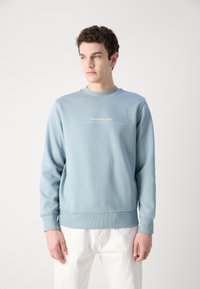 Gaiši zils džemperis, kas izgatavots no mīksta materiāla, ar apaļu kakla izgriezumu, ribotām piedurknēm un kontrastējošu logo tekstu priekšpusē.