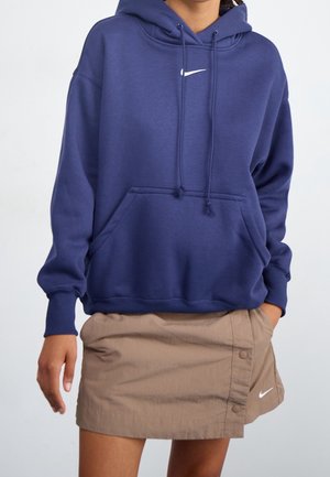 Person trägt einen dunkelblauen Nike-Hoodie mit Fronttasche und einen beigen Nike-Miniknopfrock, Hände in den Taschen des Rocks.