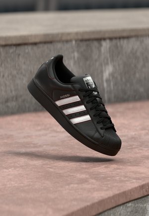 Zapatilla Adidas negra con rayas blancas y texto "Messi", mostrada en el aire sobre una superficie de hormigón rosa cerca de una pared gris.