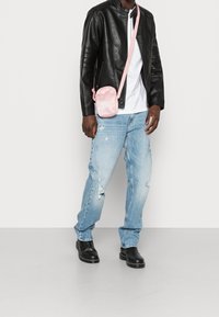 Růžová crossbody taška s logem, nošená přes černou koženou bundu, doplněná bílou košilí a světlými modrými džíny s efektem opotřebení a černými botami.