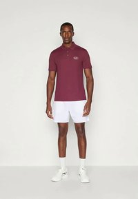 Άνδρας όρθιος που φορά μαρονο polo μπλουζάκι με το λογότυπο "EA7", λευκά σορτς, λευκές κάλτσες και λευκά αθλητικά παπούτσια, σε απλή φωτεινή φόντο.