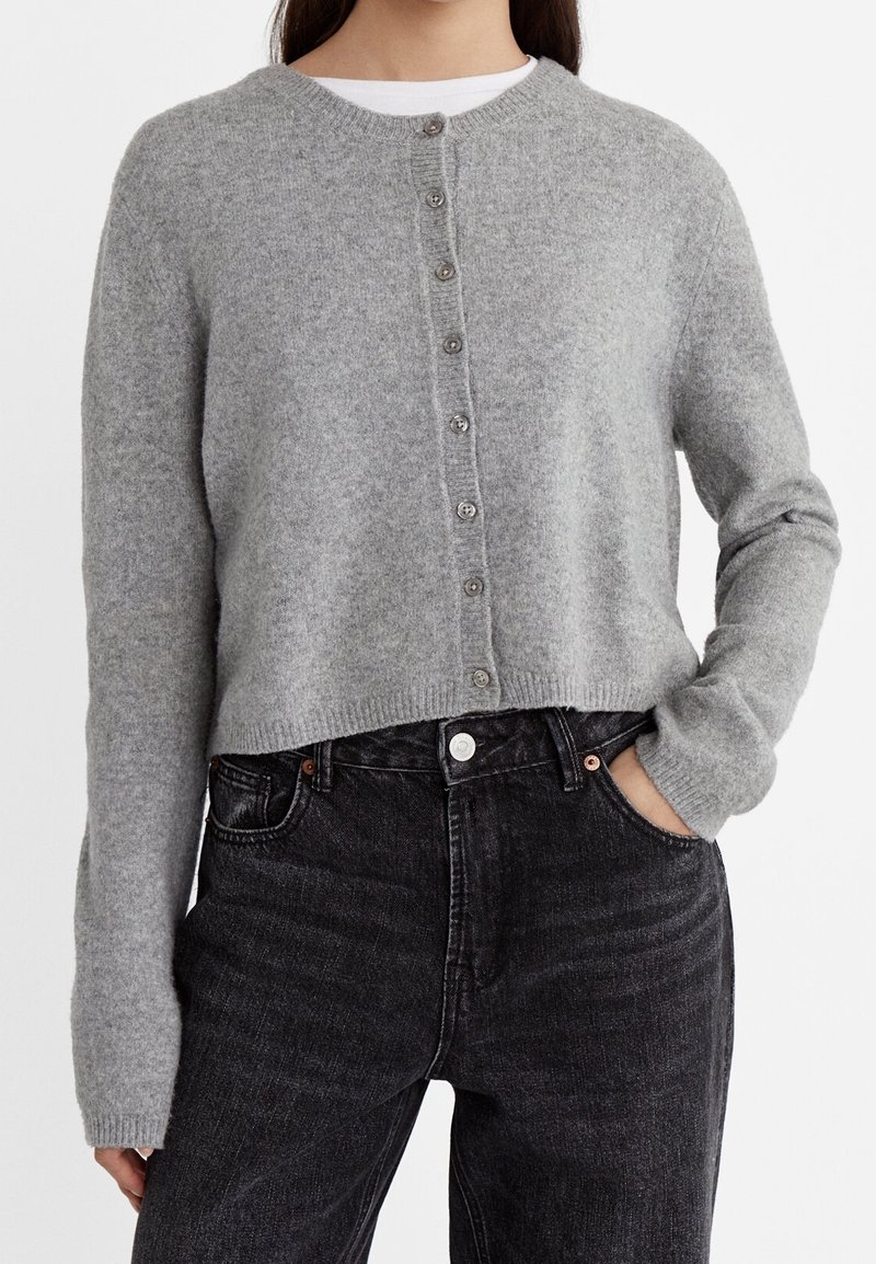 Femme portant un cardigan en laine gris clair boutonné sur une chemise blanche, associé à un jean noir taille haute, main dans la poche.