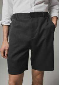 Pantaloni scurți de culoare gri închis, dintr-un material texturat, cu un croi drept, buzunare laterale și un tiv simplificat. Asortați cu o cămașă albă.