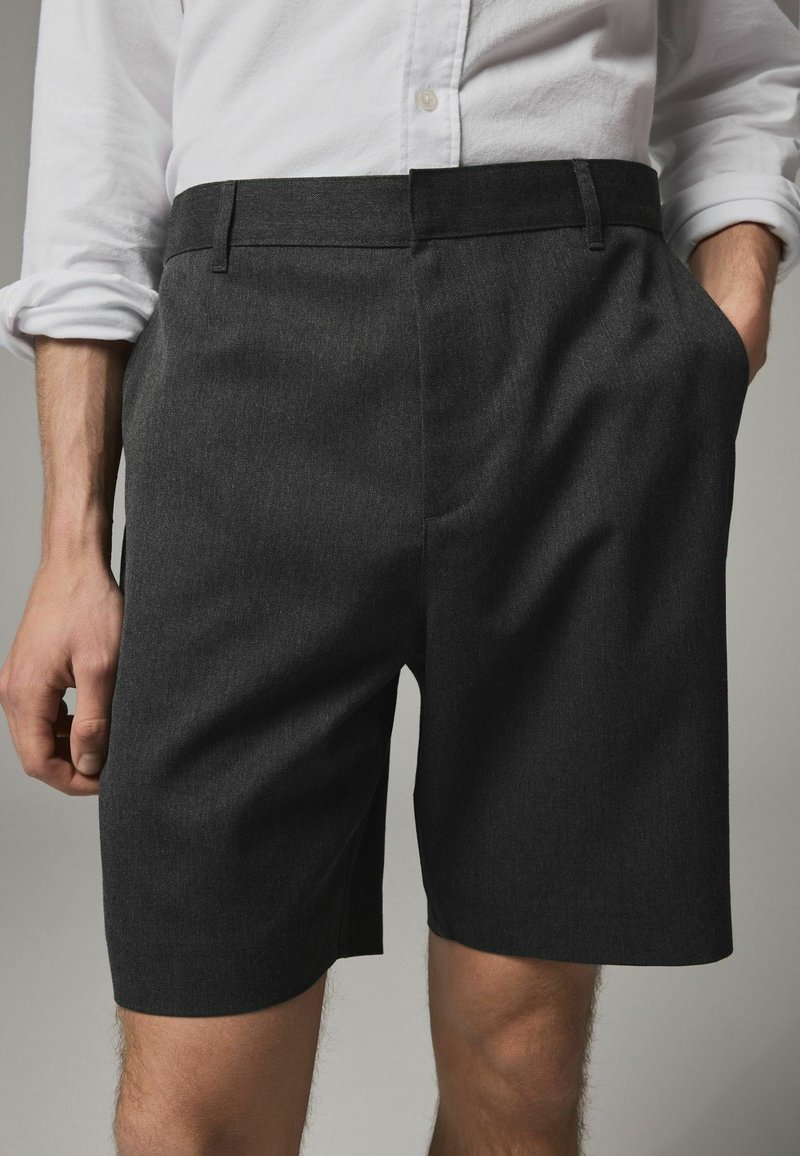 Pantaloni scurți de culoare gri închis, dintr-un material texturat, cu un croi drept, buzunare laterale și un tiv simplificat. Asortați cu o cămașă albă.