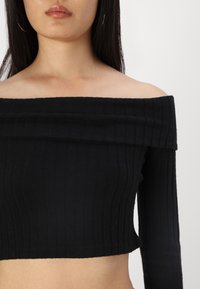 Juodas off-the-shoulder, banguotas trumpas viršus su ilgomis rankovėmis, pasižymintis vientisa tekstūra ir plačiu iškirpimu, kuris suteikia dizainui išraiškingumo.