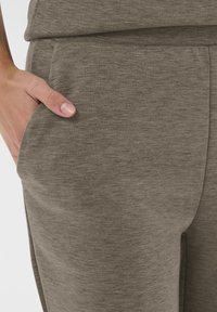 Ľavá ruka odpočíva vo vrecku nohavíc z mäkkej látky v taupe farbe s viditeľným švom a priliehavým pásom.