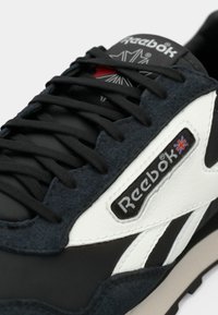 Sneaker Reebok realizzato in materiale sintetico nero, con dettagli in suede grigio scuro, logo in pelle bianca e suola in gomma testurizzata con lacci neri.