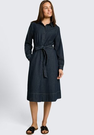 Frau trägt ein langärmeliges, geknöpftes, dunkles Denim-Midikleid mit Taillengürtel und schwarzen Slides-Sandalen, steht und schaut zur Seite.