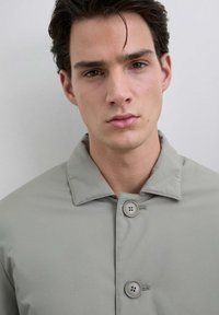 Joven con cabello oscuro, expresión neutral, vistiendo un abrigo claro de botones grises sobre un fondo claro y liso.